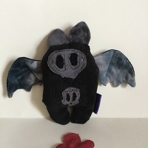 Puede incluir: Un peluche de murci&eacute;lago de pana negro con detalles grises, incluyendo orejas, hocico y alas. Las alas tienen un patr&oacute;n de te&ntilde;ido anudado azul y gris. Una flor roja con un tallo verde est&aacute; en primer plano.