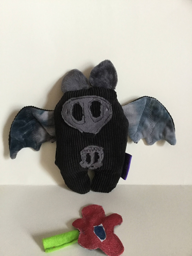 Puede incluir: Un peluche de murci&eacute;lago de pana negro con detalles grises, incluyendo orejas, hocico y alas. Las alas tienen un patr&oacute;n de te&ntilde;ido anudado azul y gris. Una flor roja con un tallo verde est&aacute; en primer plano.