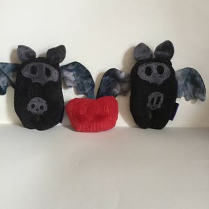 Puede incluir: Dos peluches de murci&eacute;lago de pana negra con detalles grises y alas te&ntilde;idas con tinte anudado se muestran junto a un peluche rojo en forma de coraz&oacute;n de pana. Los murci&eacute;lagos tienen orejas y caras grises, con detalles cosidos. Los juguetes est&aacute;n sobre una superficie blanca.