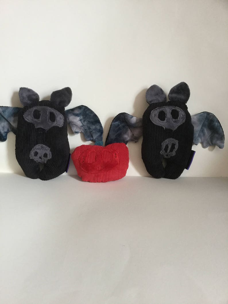 Puede incluir: Dos peluches de murci&eacute;lago de pana negra con detalles grises y alas te&ntilde;idas con tinte anudado se muestran junto a un peluche rojo en forma de coraz&oacute;n de pana. Los murci&eacute;lagos tienen orejas y caras grises, con detalles cosidos. Los juguetes est&aacute;n sobre una superficie blanca.