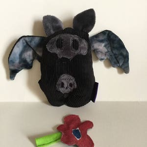 Puede incluir: Un murci&eacute;lago de peluche negro con detalles grises, incluyendo orejas, hocico y alas con un patr&oacute;n azul y gris. Tambi&eacute;n hay una flor de fieltro roja con un tallo verde. El murci&eacute;lago tiene una textura de pana.