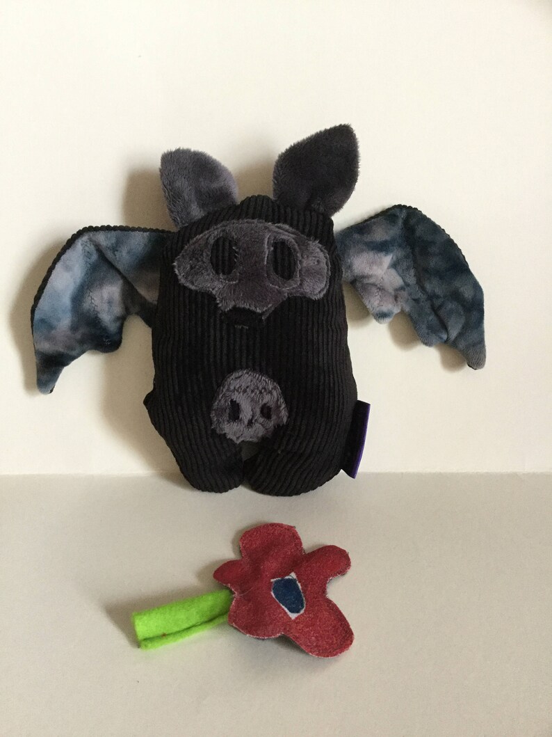 Puede incluir: Un murci&eacute;lago de peluche negro con detalles grises, incluyendo orejas, hocico y alas con un patr&oacute;n azul y gris. Tambi&eacute;n hay una flor de fieltro roja con un tallo verde. El murci&eacute;lago tiene una textura de pana.