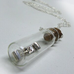 May include: A silver charm necklace with a small glass bottle pendant. The bottle contains a silver charm with the letters "RK" on it. The bottle is sealed with a cork stopper.