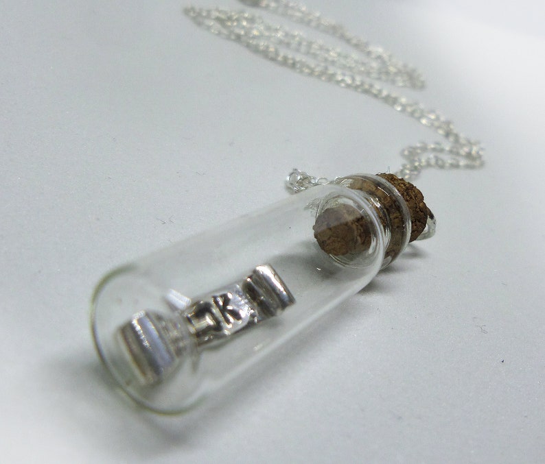 May include: A silver charm necklace with a small glass bottle pendant. The bottle contains a silver charm with the letters "RK" on it. The bottle is sealed with a cork stopper.