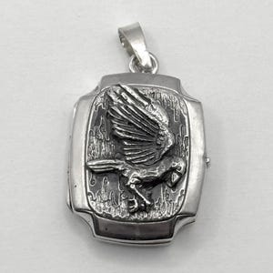 May include: A silver rectangular pendant featuring a detailed relief of a bird in flight. The bird has outstretched wings and is set against a textured background. The pendant has a loop for a chain.