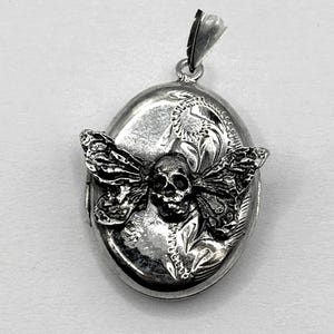 May include: Silver-toned oval pendant with a skull and butterfly wing design. The pendant features intricate floral detailing and a bail for a chain. The skull is centered, with wings extending outward.