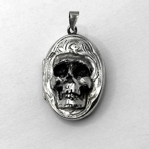 May include: Silver-toned oval locket pendant featuring a detailed skull design. The skull is centered within the locket, surrounded by an ornate floral border. The pendant has a bail for attaching to a chain.