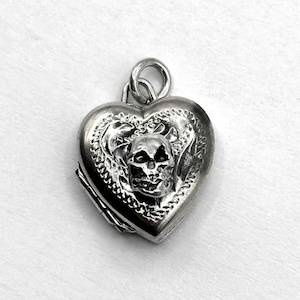May include: Silver heart-shaped pendant with a skull design. The pendant has a hinged opening and a loop for a chain. The skull is centered on the front, surrounded by a decorative border.