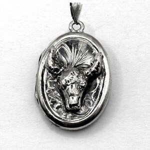 May include: Oval silver pendant featuring a detailed bull's head within an oval frame. The pendant has a loop at the top for a chain. The bull's head is in the center, with intricate detailing on the horns and face. The background has a floral design.