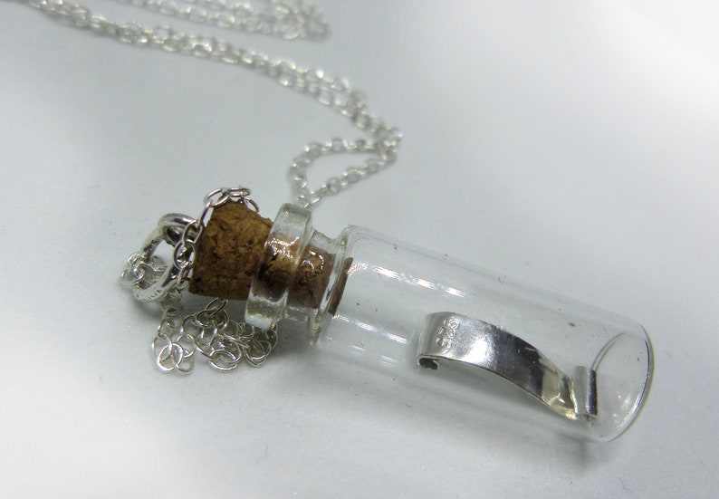 May include: A silver chain necklace with a small glass bottle pendant. The bottle has a cork stopper and contains a silver charm.