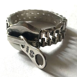Può includere: Un anello a zip in argento con una fascia testurizzata e una linguetta che dice "925".