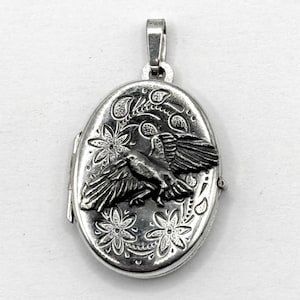May include: Silver oval locket with a bird motif. The locket showcases a raised bird with open wings, framed by floral and paisley designs. A bail is at the top, and the locket opens with a hinge. The locket is approximately 3 cm in length.