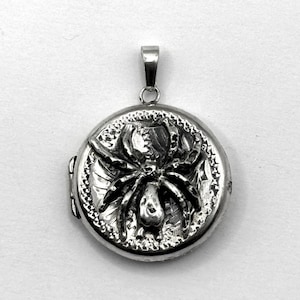 May include: A round, silver-colored locket with a detailed spider design on the front. The spider is centered, with its legs outstretched. The locket has a loop at the top for a chain. The background is plain white.