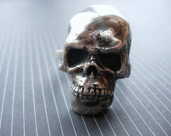 May include: A silver skull ring with a detailed design. The skull has a prominent brow, eye sockets, and a toothy grin.