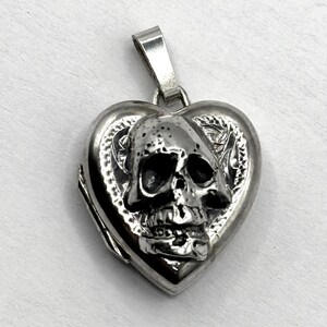 May include: A silver heart-shaped locket pendant with a skull design. The skull is centered on the front of the locket, with detailed features. The pendant has a bail for a chain.