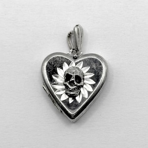 May include: Silver heart-shaped locket pendant with a skull and floral design. The locket is detailed with a skull in the center, surrounded by a floral pattern. The pendant has a bail for attaching to a chain.