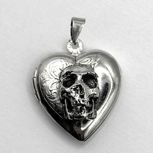 May include: A silver heart-shaped locket pendant with a skull design. The locket has an ornate floral pattern on the front and a bail for a chain. The skull is detailed and three-dimensional.