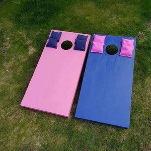 May include: Two wooden cornhole boards, one pink and one blue, with bean bags on top. The boards are on a green grassy surface.
