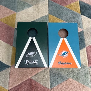 May include: Two wooden cornhole boards with team logos. The left board is green with a white triangle and the Philadelphia Eagles logo. The right board is blue with a white triangle and the Miami Dolphins logo.