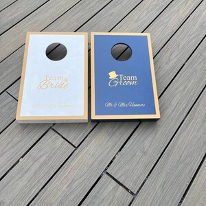 May include: Two wooden cornhole boards with a gold trim. The board on the left is white with the text "Team Bride" and "Mr. & Mrs. Hammore". The board on the right is blue with the text "Team Groom" and "Mr. & Mrs. Hammore".