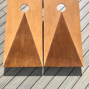 May include: Two wooden cornhole boards with a triangle design and a circular hole in the centre. The boards are stained in a light brown colour.