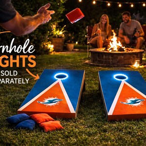 May include: Two blue and orange cornhole boards featuring a dolphin logo and a circular light ring around the hole. Four bean bags in blue and orange are on the grass. Text on the image reads: Cornhole LIGHTS SOLD SEPARATELY.