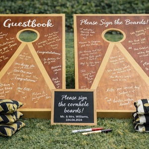 May include: Two wooden cornhole boards with the text "Guestbook" and "Please Sign the Boards!" are displayed. Each board has a hole and a triangle design. A small chalkboard reads "Please sign the cornhole boards!" with a date. Cornhole bags are stacked.