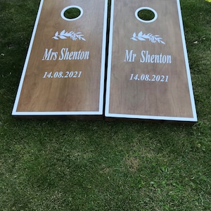May include: Two wooden cornhole boards with white trim. The boards are decorated with a white floral design and the text "Mrs. Shenton 14.08.2021" and "Mr. Shenton 14.08.2021".