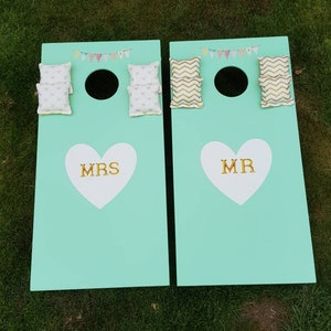 May include: Two teal green bean bag toss game boards with white hearts and gold lettering. The left board says "MRS" and the right board says "MR". Each board has three bean bags, two with a white and pink floral pattern and one with a gold chevron pattern.