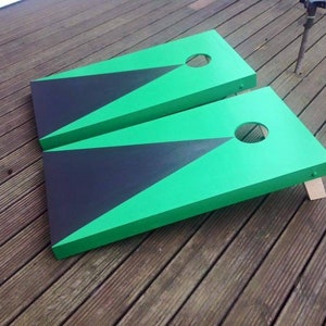 May include: Two green and black wooden cornhole boards with a hole in the centre of each board. The boards are on a wooden deck.
