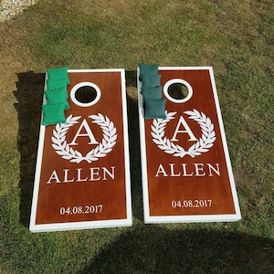 May include: Two wooden cornhole boards with white trim. Each board has a brown background with a white circle in the centre. The boards are personalised with the letter "A" inside a wreath and the name "ALLEN" below. The date "04.08.2017" is printed below the name. Each board has six green bean bags.