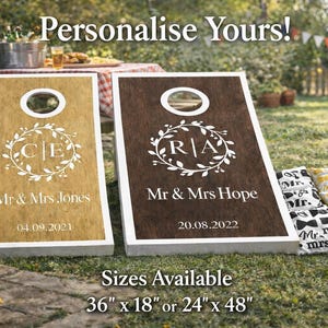 May include: Two wooden cornhole boards with white frames and circular holes. One board is light brown with 'C|E', 'Mr & Mrs Jones', and '04.09.2021'. The other is dark brown with 'R|A', 'Mr & Mrs Hope', and '20.08.2022'. Text: 'Personalise Yours!' and '36" x 18" or 24" x 48"'.
