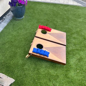 May include: Two wooden cornhole boards with a natural wood finish and a geometric triangle design. One board features four red bean bags, and the other board features four blue bean bags. Each board has a circular hole at the top centre.