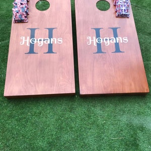May include: Two wooden cornhole boards with the name "Hogan's" printed on each. The boards are brown and have a hole in the centre. There are red, white and blue bean bags on each board.