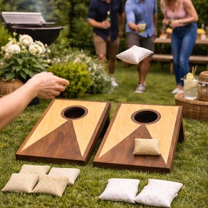 May include: Two wooden cornhole boards with a light wood and dark brown design, set on a grassy lawn. Several bean bags are scattered around the boards, and people are playing the game in the background.
