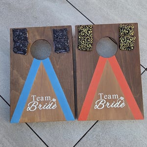 May include: Two wooden cornhole boards with a brown stain. One board has a blue triangle and the text "Team Bride". The other board has an orange triangle and the same text. Each board has two fabric pieces with decorative patterns.