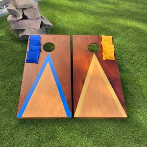 May include: Two rectangular wooden cornhole boards with a dark stained finish. One board features a large blue triangle design with four blue bean bags, while the other board has a yellow triangle design with four yellow bean bags.