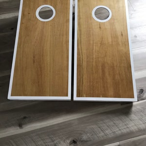 May include: Two wooden cornhole boards with a light brown wood grain finish. Each board has a white border and a white-rimmed hole near the top. The boards are designed for the game of cornhole.