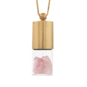 Puede incluir: Un collar de cadena dorada con un colgante que presenta un vial de vidrio transparente lleno de cristales de cuarzo rosa. El vial está coronado con una tapa dorada.