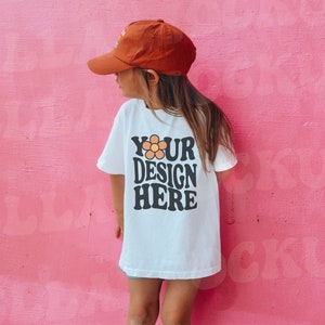 Puede incluir: Una persona joven con una camiseta blanca con el texto "YOUR DESIGN HERE" y una gorra de b&eacute;isbol marr&oacute;n. La camiseta tiene un gr&aacute;fico de una flor. La persona est&aacute; de pie frente a una pared rosa.