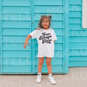 May include: A young person wearing a white t-shirt with the text "Your Design Here" in a retro font. The person is standing in front of a blue wooden wall.