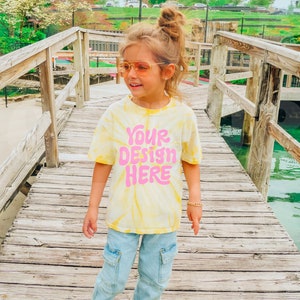 May include: A young person wearing a yellow tie-dye t-shirt with the text "YOUR DESIGN HERE" in pink. The person is standing on a wooden boardwalk, wearing light blue jeans and sunglasses.