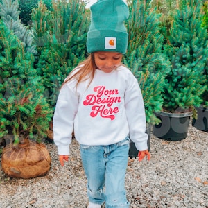 May include: A young person wearing a white sweatshirt with the text "Your Design Here" in pink lettering. They are wearing a green beanie and blue jeans and standing in front of a row of potted evergreen trees.