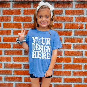 May include: A young person wearing a blue t-shirt with the text "YOUR DESIGN HERE" in white. The person is making a peace sign with their right hand and is standing in front of a brick wall.