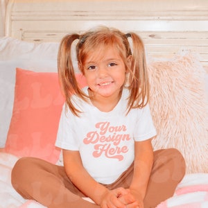 May include: A young girl wearing a white t-shirt with the text "Your Design Here" in pink. She is sitting on a bed with pink and white bedding and pillows.