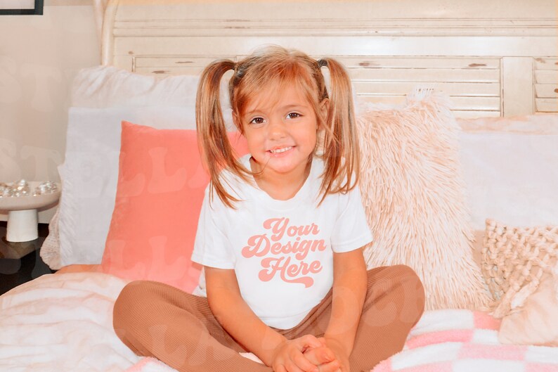May include: A young girl wearing a white t-shirt with the text "Your Design Here" in pink. She is sitting on a bed with pink and white bedding and pillows.