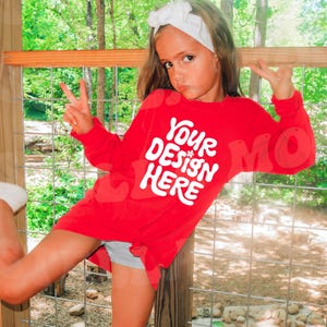 Peut inclure: Une jeune personne portant un t-shirt rouge à manches longues avec le texte "YOUR DESIGN HERE" en blanc. La personne est debout devant une clôture en bois avec un fond vert.