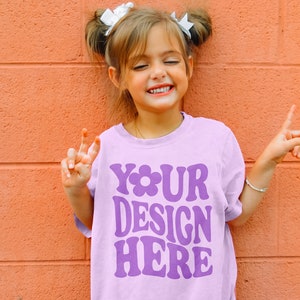May include: A young girl wearing a purple t-shirt with the text "YOUR DESIGN HERE" in a retro font. She is smiling and making peace signs with her hands.