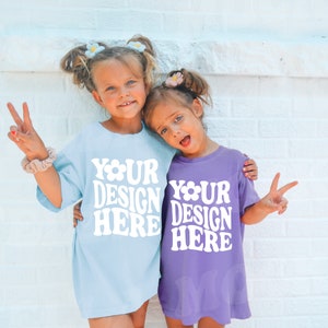 May include: Two children wearing light blue and purple t-shirts with the text "Your Design Here" printed on them. They are smiling and making peace signs with their hands.