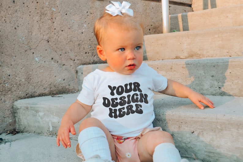 May include: A white baby onesie with black text that reads "YOUR DESIGN HERE" in a retro font. The onesie is being worn by a baby sitting on concrete steps.